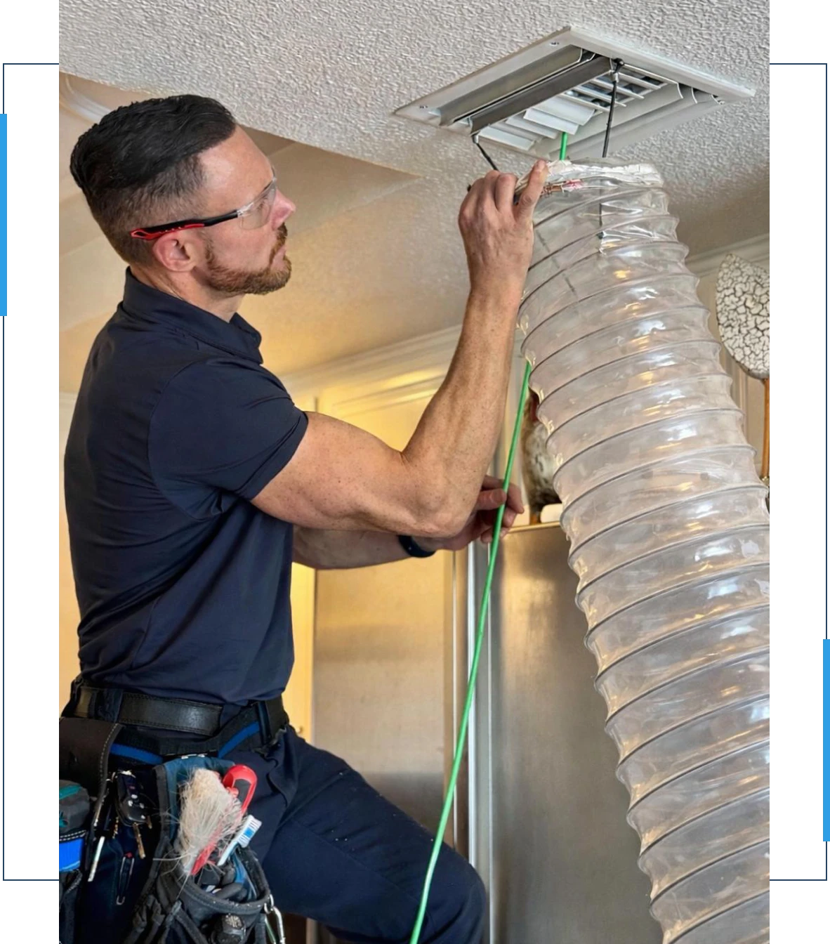 A man wearing safety glasses and a tool belt cleans or repairs a ceiling vent using a flexible hose and a green tool or cable.